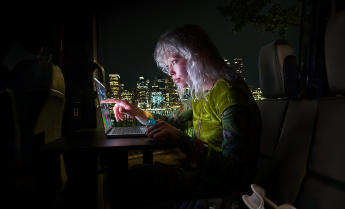 A person sitting in the back seat of a van, the open passenger door reveals a city skyline at night, MacBook Pro with PROLED XDR Display on a worktable, the person is illuminated by the screen while using the Touch feature to edit a video in DaVinci Resolve Studio